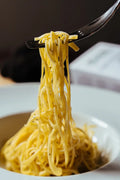 Forchetta che solleva tagliolini da un piatto bianco, con piccoli frammenti scuri visibili nella pasta