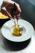 Mani che dispongono un nido di tagliolini in un piatto bianco usando una pinza e un mestolo